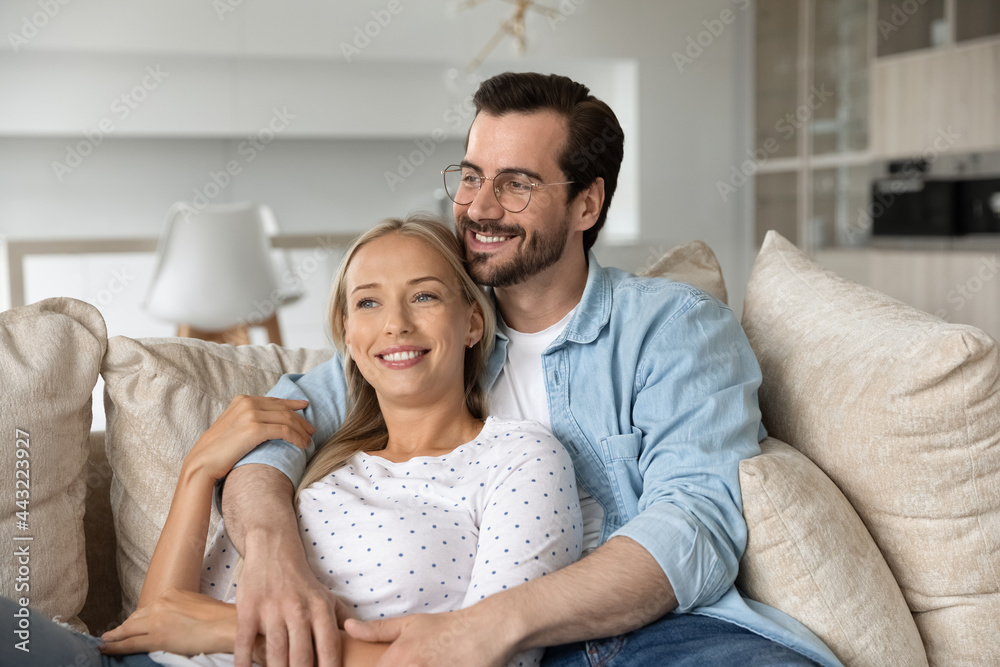 Dreamy happy young couple hugging, relaxing on couch together, smiling woman and man in glasses looking to aside in distance, visualizing good future planning, enjoying lazy leisure time on sofa - obrazy, fototapety, plakaty 