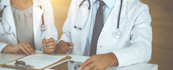 Two unknown doctors discussing treatment problems while sitting at the desk in sunny hospital office, close-up