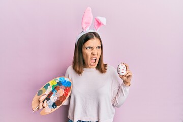 Sticker - Young beautiful woman wearing cute easter bunny ears holding painting palette and egg angry and mad screaming frustrated and furious, shouting with anger. rage and aggressive concept.