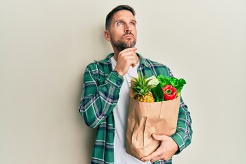 Sticker - Handsome man with beard holding paper bag with groceries with hand on chin thinking about question, pensive expression. smiling with thoughtful face. doubt concept.