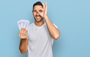 Wall Mural - Handsome man with beard holding 20 swedish krona banknotes smiling happy doing ok sign with hand on eye looking through fingers