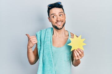 Poster - Young hispanic man wearing summer style holding yellow sun pointing thumb up to the side smiling happy with open mouth