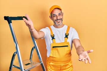 Poster - Handsome middle age man with grey hair holding ladder looking at the camera smiling with open arms for hug. cheerful expression embracing happiness.