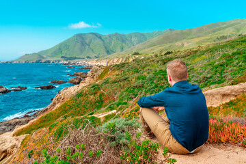 A young man is sitting on a high slope with a panoramic view of the picturesque ocean and mountains in California. Coast along the Pacific Coast Highway