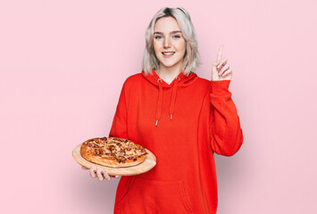 Sticker - Young blonde girl holding italian pizza surprised with an idea or question pointing finger with happy face, number one