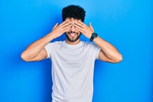 Young Arab Man With Beard Wearing Casual White T Shirt Covering Eyes With Hands Smiling Cheerful And Funny. Blind Concept.