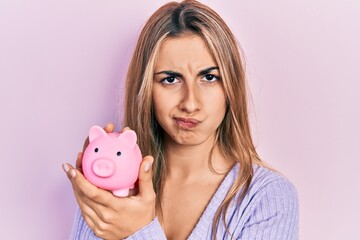 Sticker - Beautiful hispanic woman holding piggy bank skeptic and nervous, frowning upset because of problem. negative person.