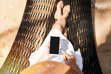 Young woman traveler lying on a hammock and using smartphone at the beach while traveling for summer vacation