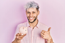 Young Hispanic Man With Modern Dyed Hair Holding Piggy Bank Smiling Happy And Positive, Thumb Up Doing Excellent And Approval Sign