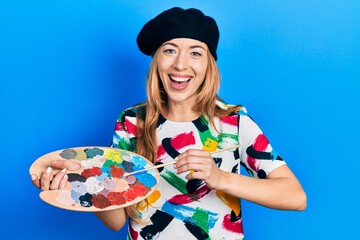 Wall Mural - Young caucasian woman artist holding paintbrush and palette smiling and laughing hard out loud because funny crazy joke.