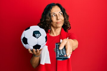 Sticker - Middle age hispanic woman football hooligan holding ball using tv control smiling looking to the side and staring away thinking.