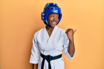 Canvas Print - Young african american girl wearing taekwondo kimono and protection helmet pointing thumb up to the side smiling happy with open mouth