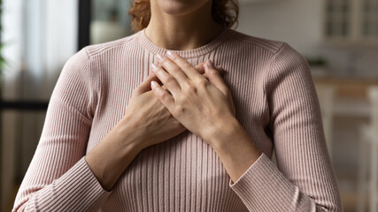 Close up cropped young woman holding folded hands on heart chest, feeling grateful. Sincere kind female volunteer showing care and devotion gesture, expressing gratitude indoors, charity concept.