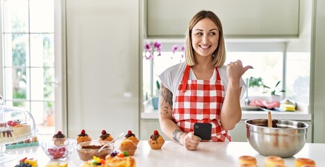 Canvas Print - Young beautiful blonde woman wearing apron cooking pastries looking for recipe on smartphone pointing thumb up to the side smiling happy with open mouth