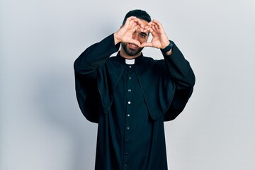 Canvas Print - Handsome hispanic man with beard wearing catholic priest robe doing heart shape with hand and fingers smiling looking through sign