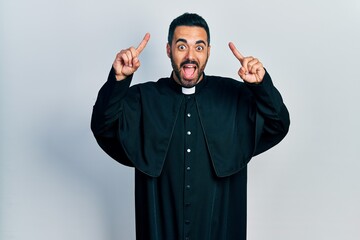 Canvas Print - Handsome hispanic man with beard wearing catholic priest robe smiling amazed and surprised and pointing up with fingers and raised arms.