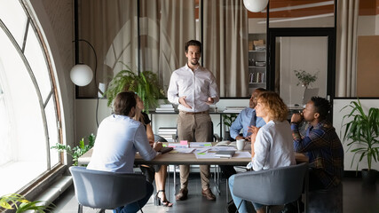 Poster - Confident businessman mentor leading briefing with diverse employees sitting at table in office boardroom, business coach leader training staff, explaining marketing plan or project strategy