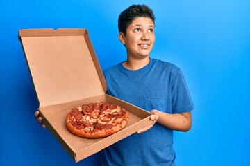 Teenager hispanic boy eating tasty pepperoni pizza smiling looking to the side and staring away thinking.