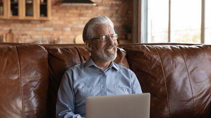 Wall Mural - Happy mature senior 70s man using laptop, sitting on sofa, learning online or working from home, looking away in good thoughts, thinking over webinar, training, workshop