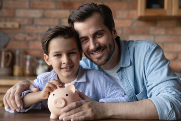 Happy dad and little son holding piggy bank, smiling, looking at camera. Father and kid saving money together for education, safe wealthy future, keeping good financial management. Head shot portrait