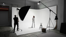 Fashion Photography In A Photo Studio. Professional Male Photographer Taking Pictures Of Beautiful Woman Model On Camera, Backstage