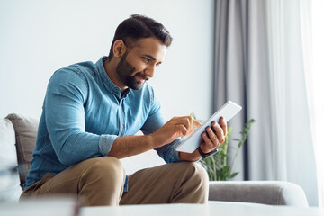 Millennial indian man using tablet reading news browsing internet, watching videos, checking social networks relaxing on couch sofa in living room. Online work, education or communication concept
