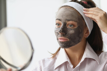 Wall Mural - Young woman with black clay mask looking in mirror at home