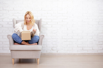 Wall Mural - young beautiful woman sitting in armchair and opening parcel box with her order - copy space over white brick wall