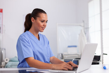 medical nurse in uniform using laptop sitting at desk in hospital office. health care physician usin