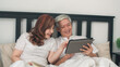 © tirachard - Asian senior couple using tablet at home. Asian Senior Chinese grandparents, husband and wife happy after wake up, watching movie lying on bed in bedroom at home in the morning concept.