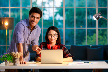 Male and female couple staying in living room together at evening twilight and working with notebook computer, looking to camera with happy faces. New normal, work at home concept