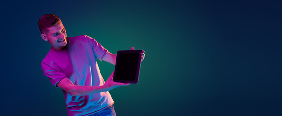 Young Caucasian man with digital tablet isolated over gradient blue green studio background in pink neon light with copyspace for ad. Concept of human emotions