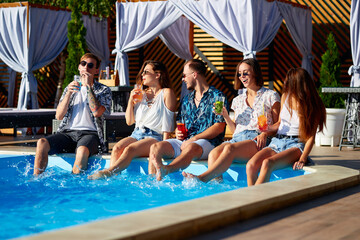 Group of friends having fun at poolside party clinking glasses with fresh cocktails sitting by swimming pool on sunny summer day. People toast drinking beverages at luxury villa on tropical vacation.