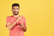 © khosrork - Smiling positive male gamer with beard in striped t-shirt holding gamepad pointing finger at empty space, advertising of video games competition. Indoor studio shot isolated on yellow background