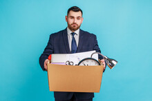 Portrait Of Sad Depressed Man Employee In Suit Standing And Holding In Hands Things In Cardboard Box, Feeling Sadness, Dismissed From Job. Indoor Studio Shot Isolated On Blue Background.