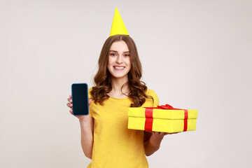 Wall Mural - Birthday present, bonus for mobile user. Portrait of happy teenager girl in birhday cone holding gift box and cell phone with mock up, blank display. Indoor studio shot isolated on gray background.