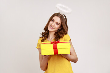 Poster - Cheerful young teenage girl in casual yellow T-shirt with pleasant smile and halo overhead holding gift box, charity organization, religion. Indoor studio shot isolated on gray background.