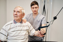 Confused Aging Man Pulling Stretch Strap With Help Of Doctor