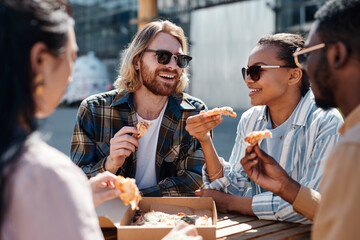 Diverse group of carefree young people enjoying pizza outdoors, scene lit by sunlight, copy space