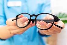 Doctor Optometrist In Blue Uniform Holds In Her Hands And Gives A Pair Of Black Rimmed Glasses To A Patient In A Clinic, Optical Store. Ophthalmology, Vision Correction And Protection, Eye Health