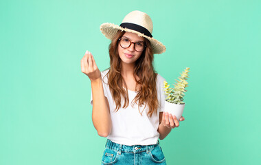 young pretty woman making capice or money gesture, telling you to pay with a straw hat and holding a cactus