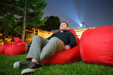 Handsome man laying on pillow on grass and watching movie at outdoor cinema in public park. Perfect spending evening or weekend time in open air. Leisure and entertainment for people at summer.