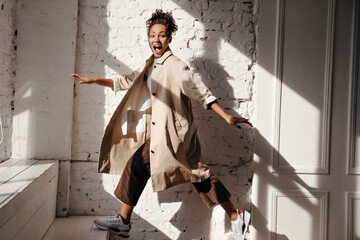 Amazed black young woman in trench coat looking at camera. Full length view of african american model jumping with smile.