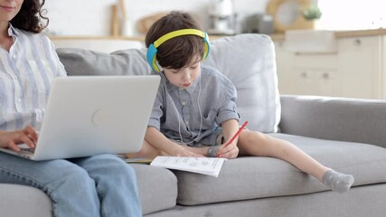 Wall Mural - Home education: small kid listen lesson wearing headphones and write in copybook sitting next to mom work online on laptop on freelance or remote occupation. Cute preschool boy learning homeschooling