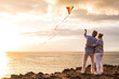 © Daniel - close up and portrait of two old and mature people playing and enjoying with a flaying kite at the beach with the sea at the background with sunset - active seniors having fun.