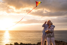 Close Up And Portrait Of Two Old And Mature People Playing And Enjoying With A Flaying Kite At The Beach With The Sea At The Background With Sunset - Active Seniors Having Fun.