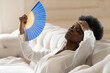 Tired Afro business woman suffering from heatstroke or hot summer flat without air-conditioner, touching her forehead, using waving fan, resting, sitting in living room at home. Heat concept