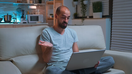 Poster - Focused man sitting on sofa in pajamas while talking with collegue during videocall conference using laptop computer. Caucasian male working at communication project late at night in kitchen