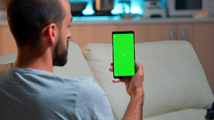 Poster - Relaxed man looking at smartphone with mock up green screen chroma key display while relaxing on sofa. Caucasian male holding in horizontal mode phone with isolated display late at night in kitchen