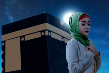 Wall Mural - Asian Muslim woman in a veil standing and praying with Kaaba view